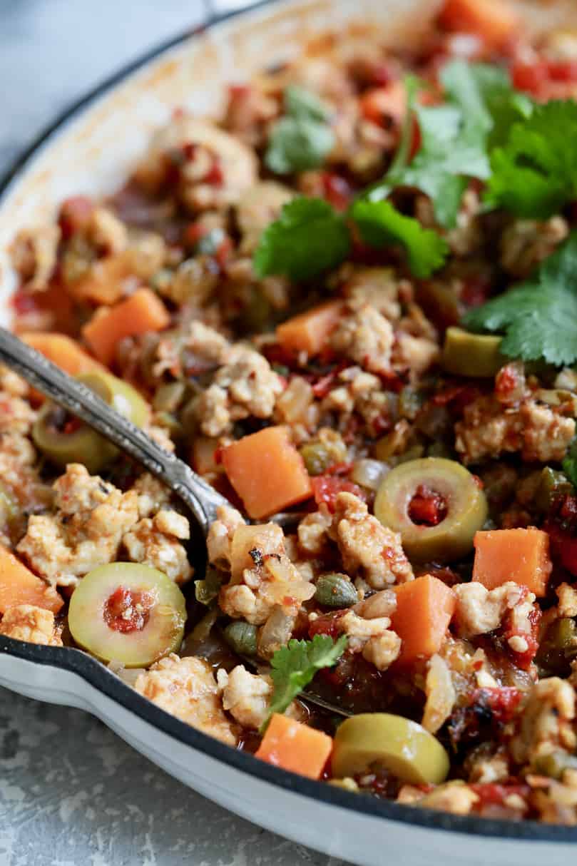 Close-up photo of Cuban-Style Turkey Picadillo with Sweet Potatoes in white skillet with spoon about to remove some.