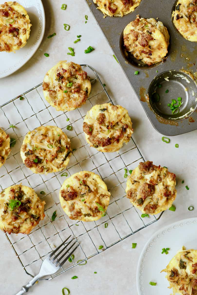 Overhead photo of Hash Brown Breakfast Muffins with Poblano Peppers and Gouda Cheese on cooling rack with some still in muffin pan and some on plates.
