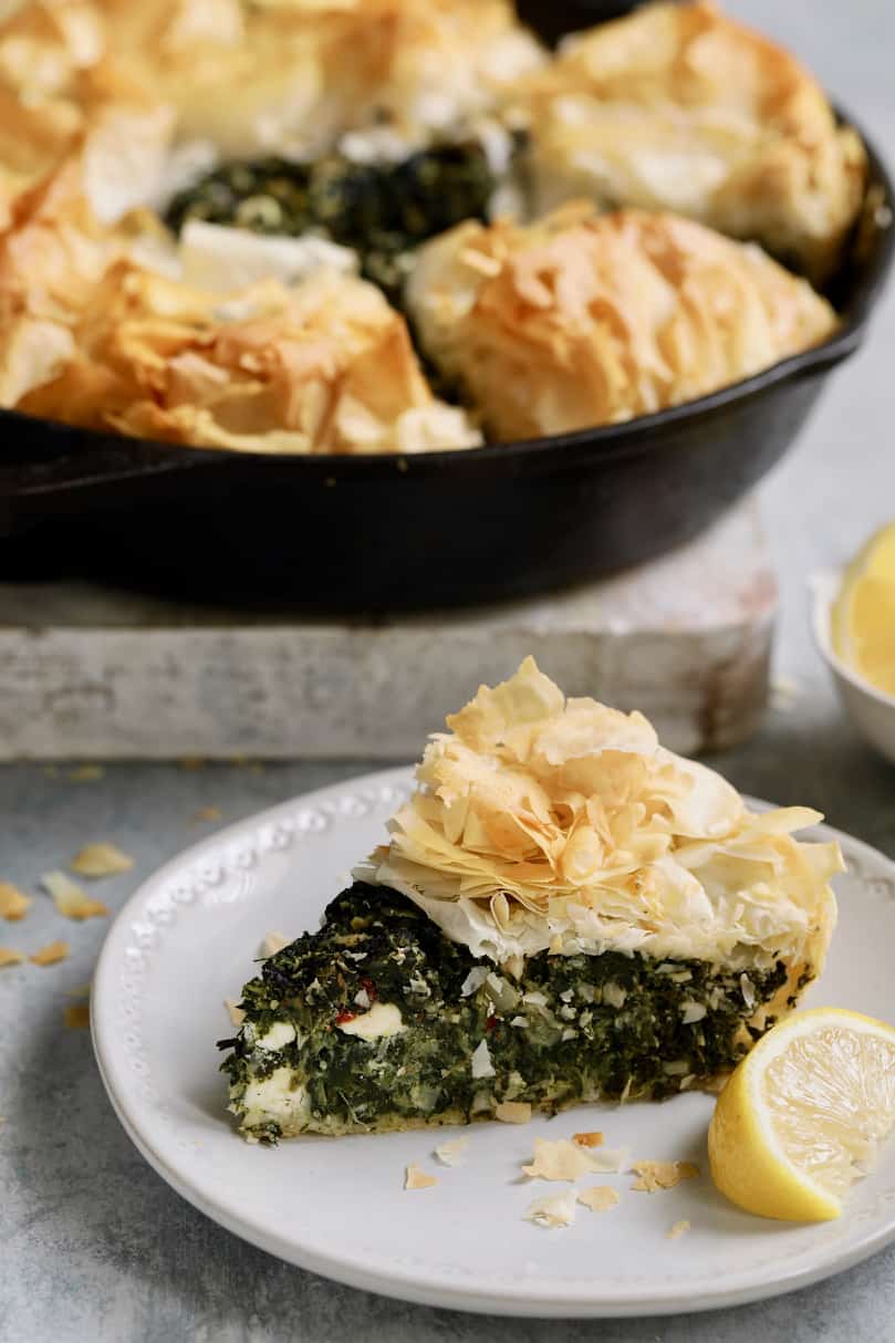Photo of one slice of Skillet Spanakopita (Greek Spinach and Feta Pie) on small white plate with rest of the pie in the background.