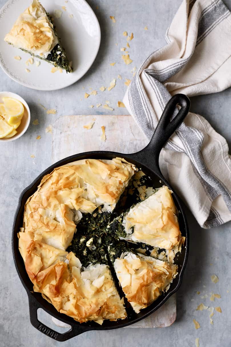Photo of baked spanakopita on light gray background with a slice cut and placed on a white plate.