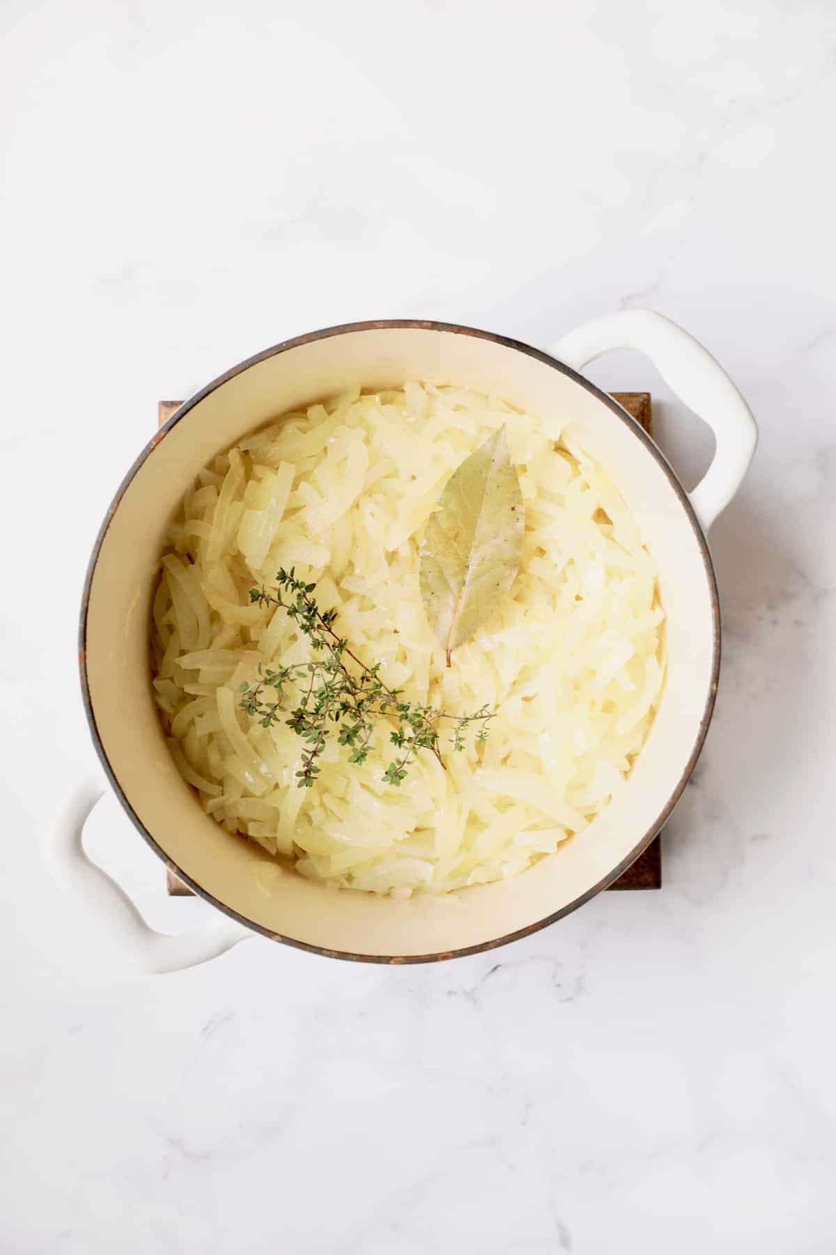 Overhead shot of partially cooked onions with thyme and bay leaf in white Dutch oven