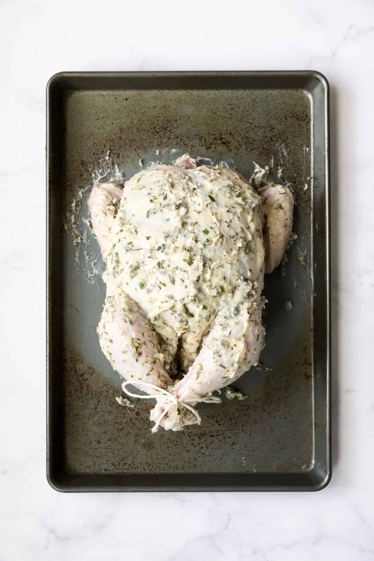 Photo of uncooked whole chicken rubbed with butter on baking sheet.