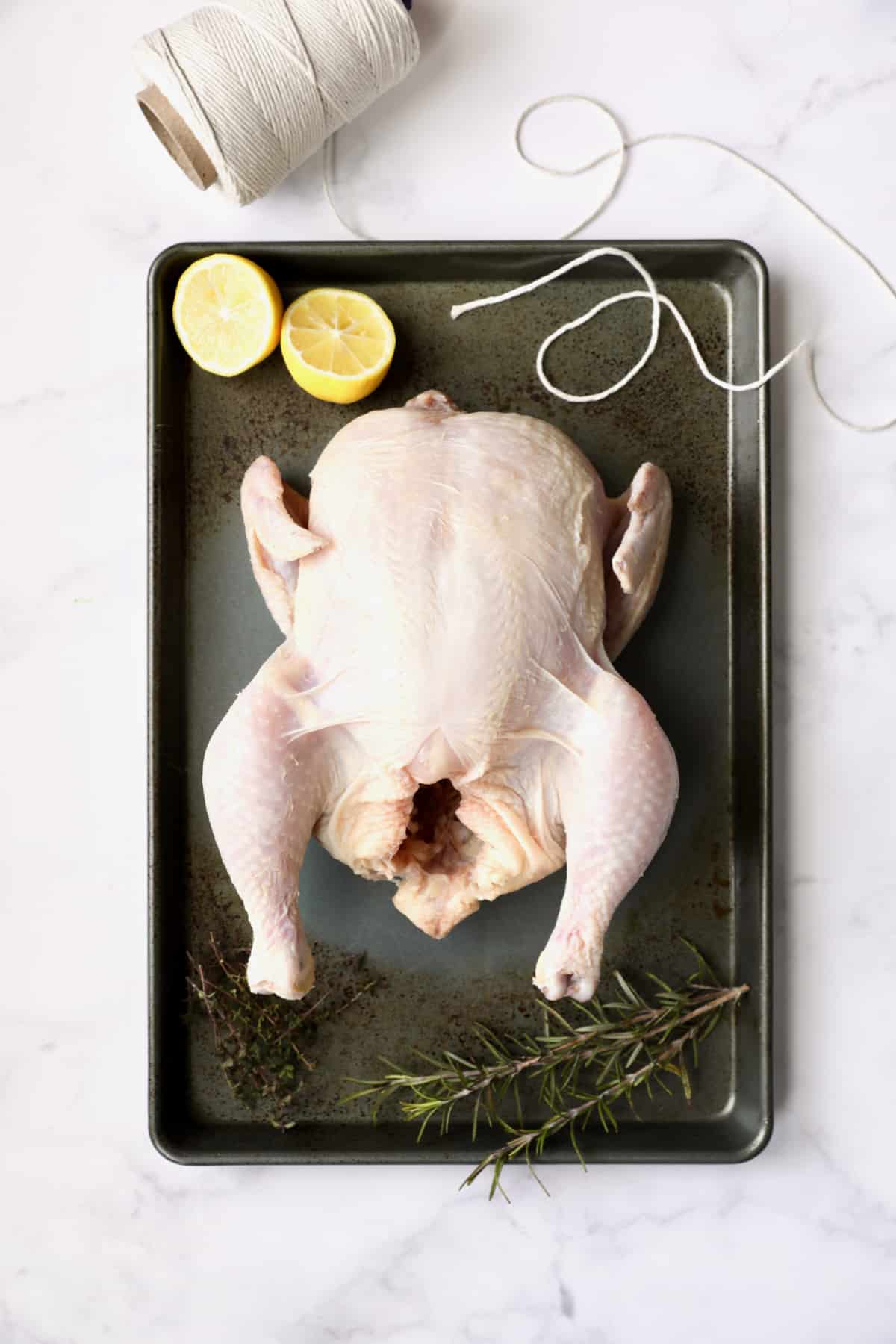 Photo of uncooked whole chicken on baking sheet with butcher's twine, lemon halves and herb sprigs.