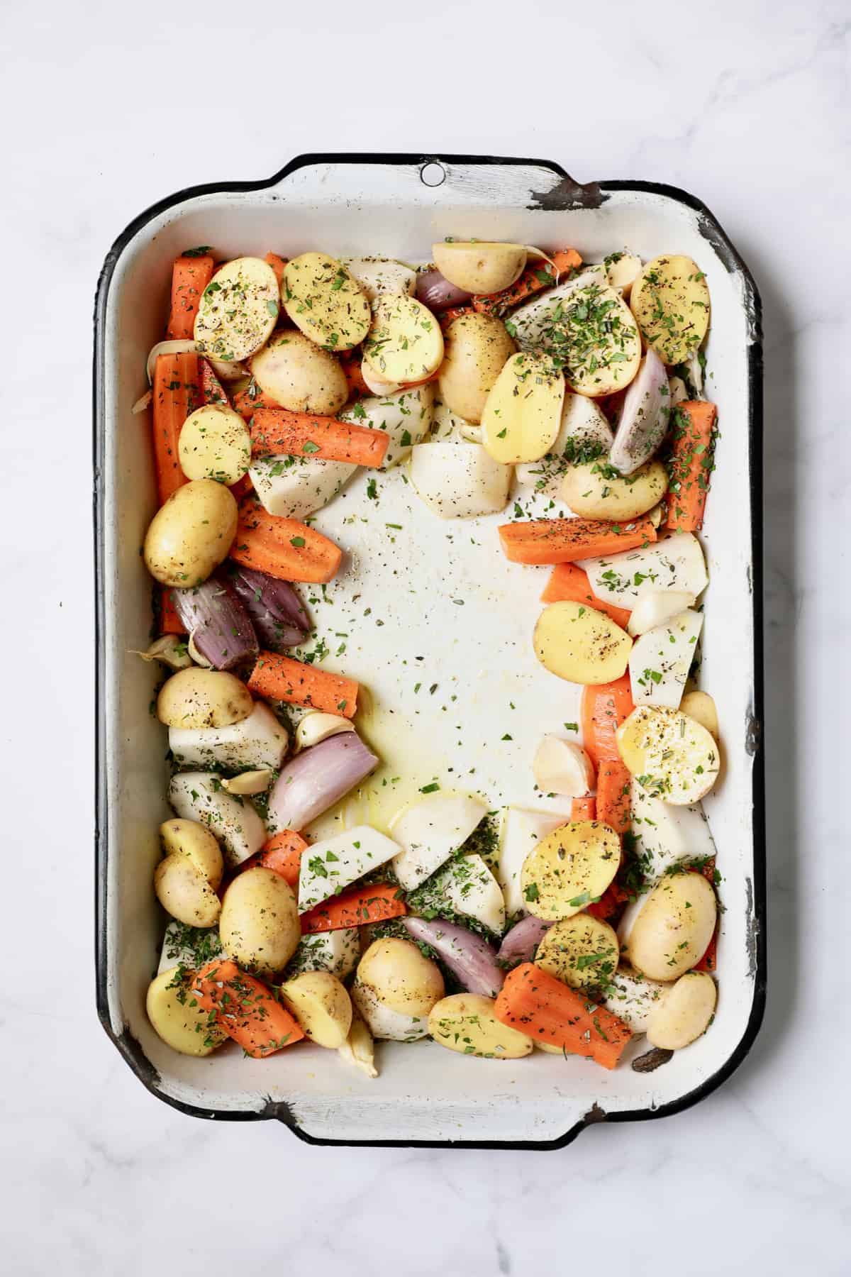Photo of uncooked root vegetables in white roasting pan sprinkled with fresh herbs.