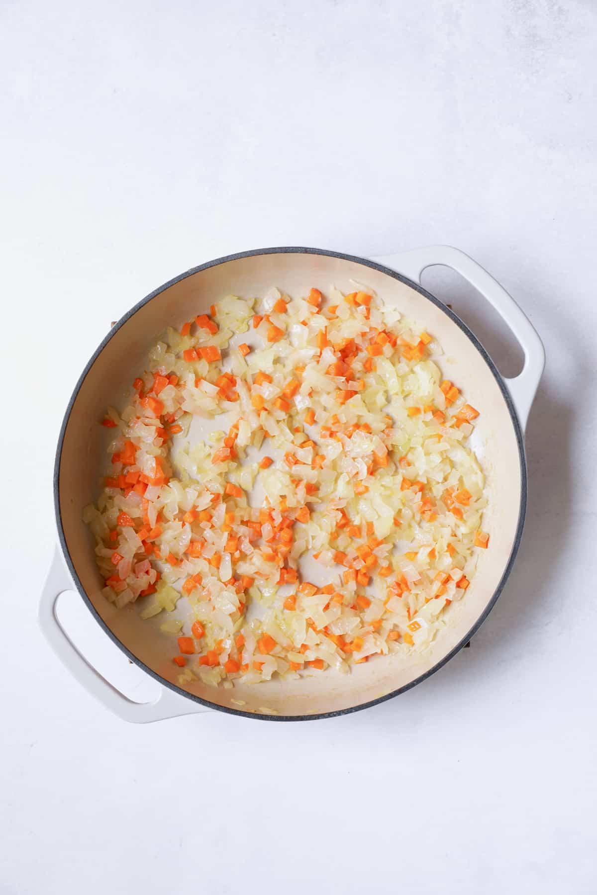 Photo of onion and carrot being cooked in white cast iron pan.