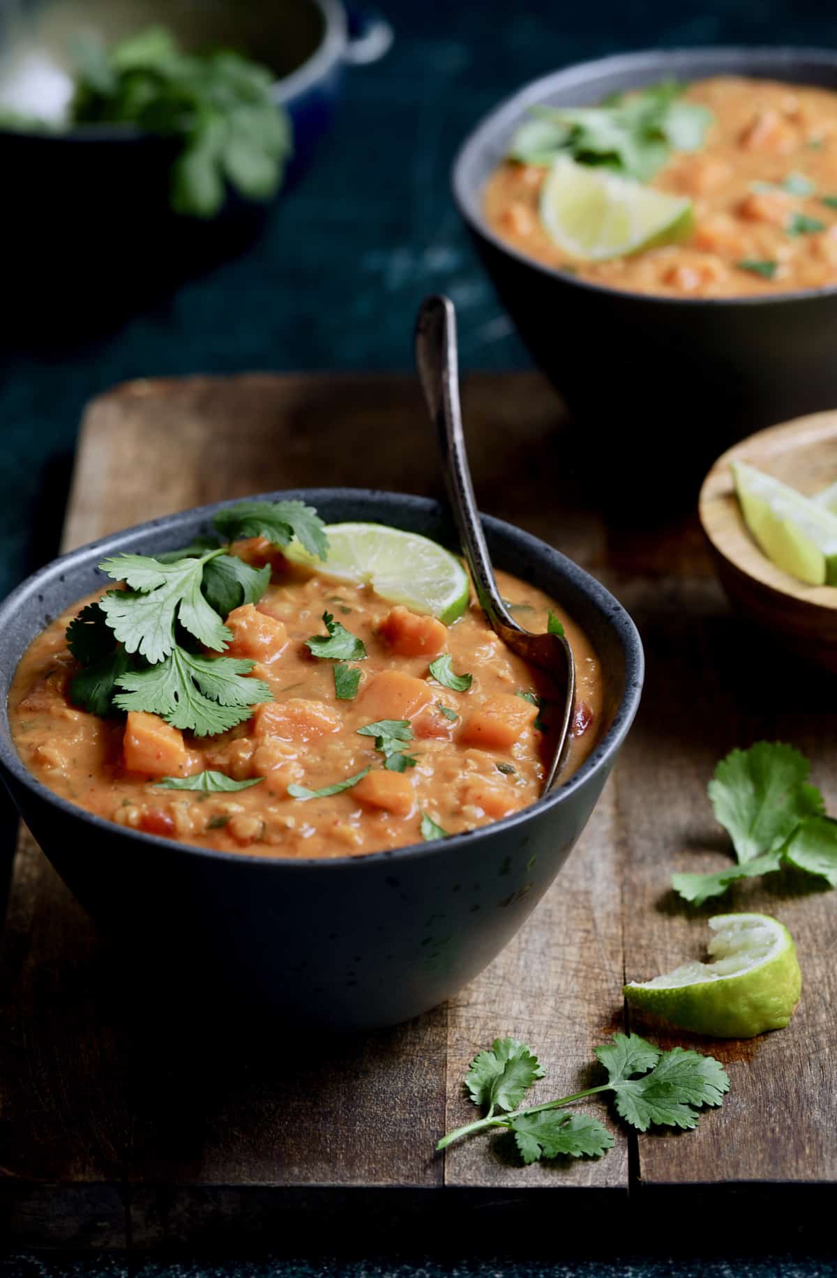Photo of Thai Red Curry Sweet Potato and Lentil Soup in gray bowl on wood surface garnished with lime and cilantro.