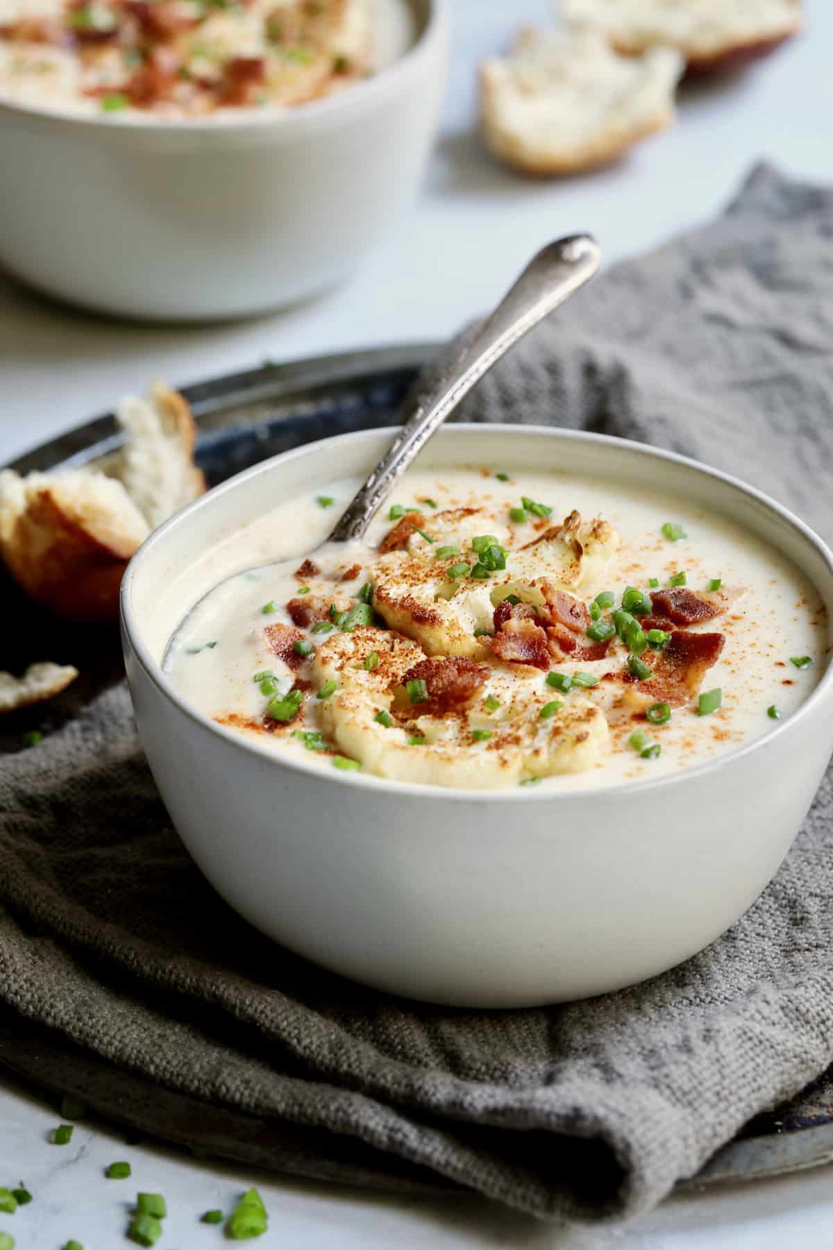 Cauliflower Potato Soup with Smoked Gouda in white bowl with soup spoon garnished with bacon, chives and cauliflower.