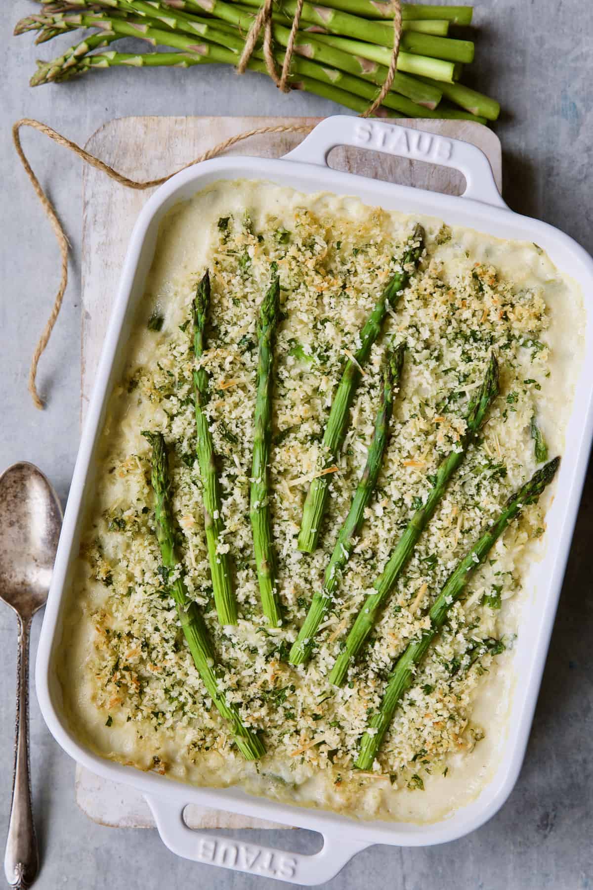 Photo of Creamy Asparagus and Rice Casserole in white baking dish with serving spoon and bunch of asparagus.