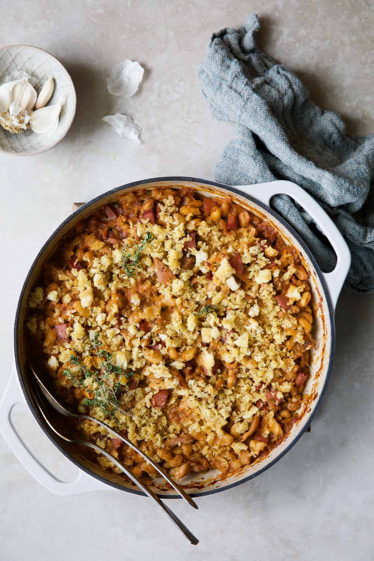 Photo of Easy Weeknight Chicken and Sausage Cassoulet with serving utensils with blue napkin and garlic cloves.