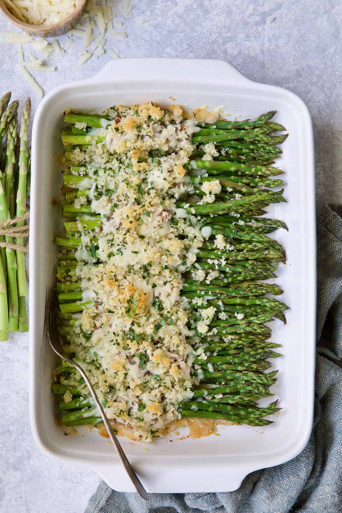 Photo of Asparagus Cordon Bleu in white rectangular baking dish with uncooked asparagus on the side and cheese scattered about.