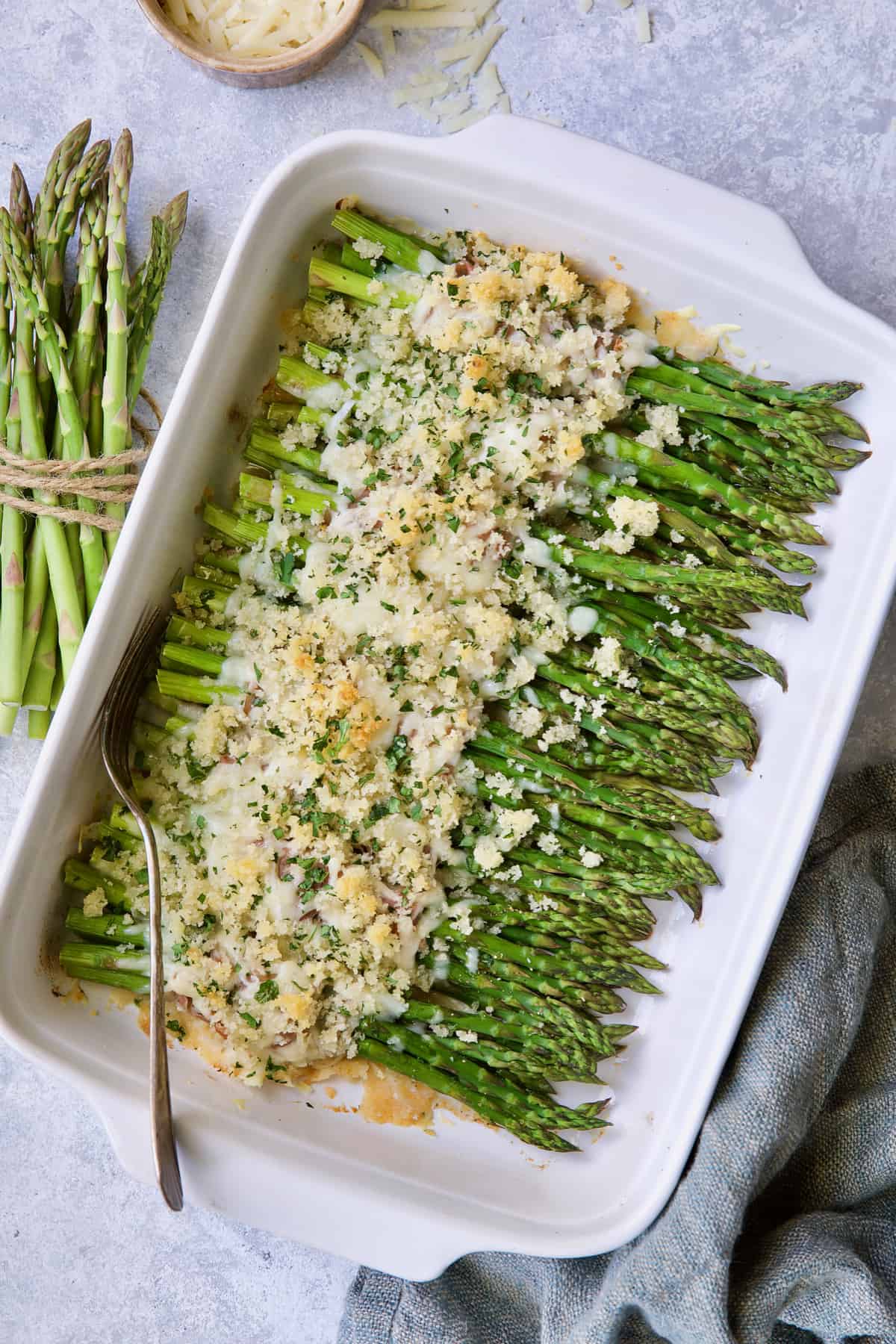 Photo of Asparagus Cordon Bleu in white baking dish on light blue background with cheese scattered about.