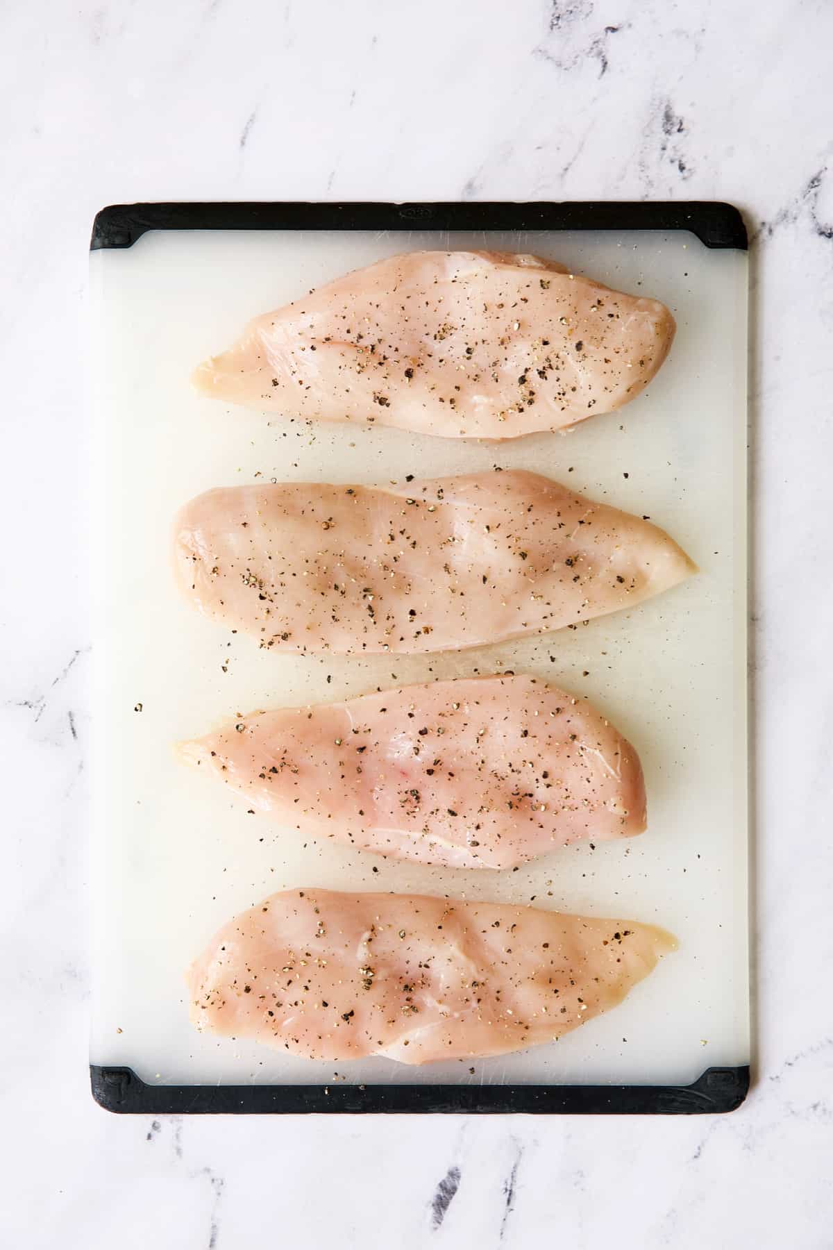 Four boneless skinless chicken breasts on a white cutting board seasoned with salt and pepper.