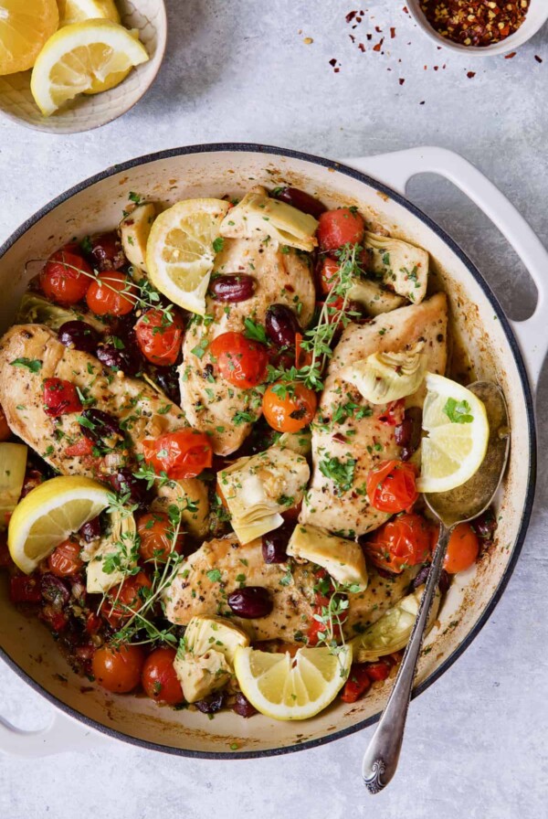 Four Mediterranean chicken breasts with tomatoes, artichokes and lemon slices in white enamel cast iron skillet.