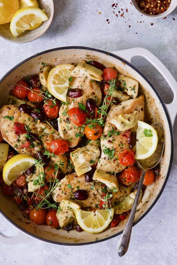 Photo of Easy Skillet Mediterranean Chicken in white enamel cast iron skillet garnished with fresh thyme and lemon wedges.