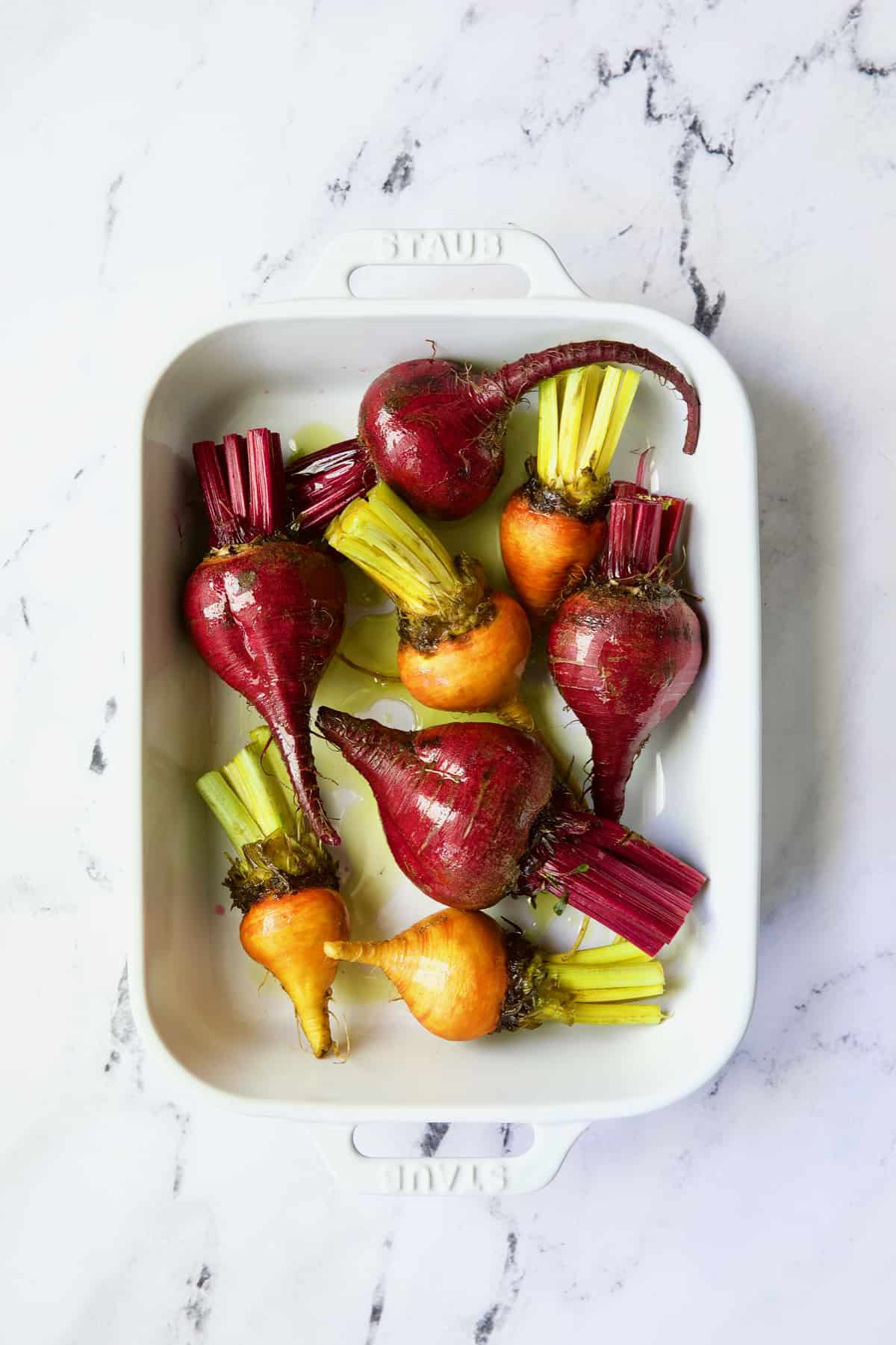 Uncooked red and yellow beets in white baking dish drizzled with oil.