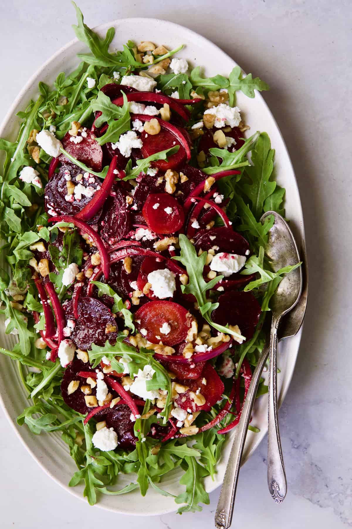 Roasted Beet Salad with Walnuts Goat Cheese and Honey Balsamic Dressing with serving utensils on white oval platter.