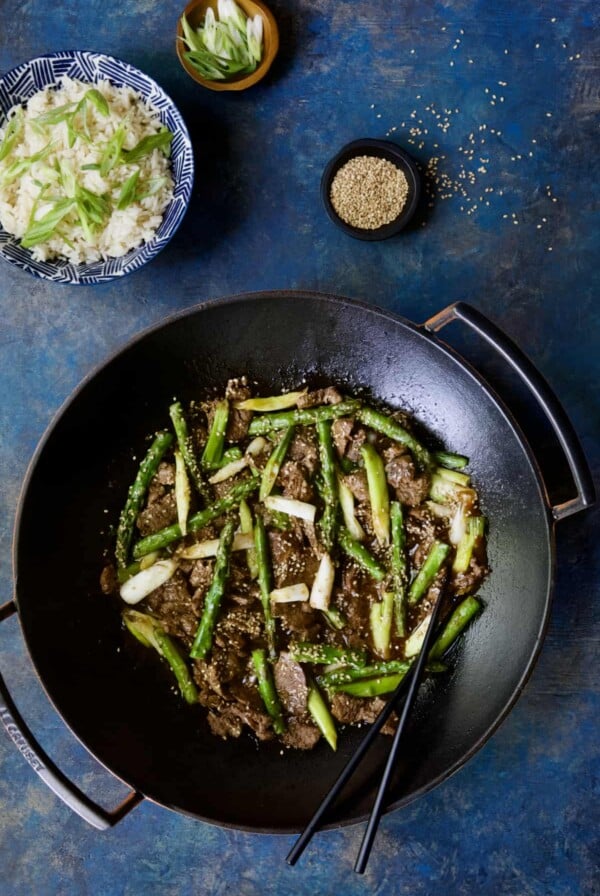 Photo of Sesame Beef and Asparagus Stir-Fry in cast iron wok on bright blue background.