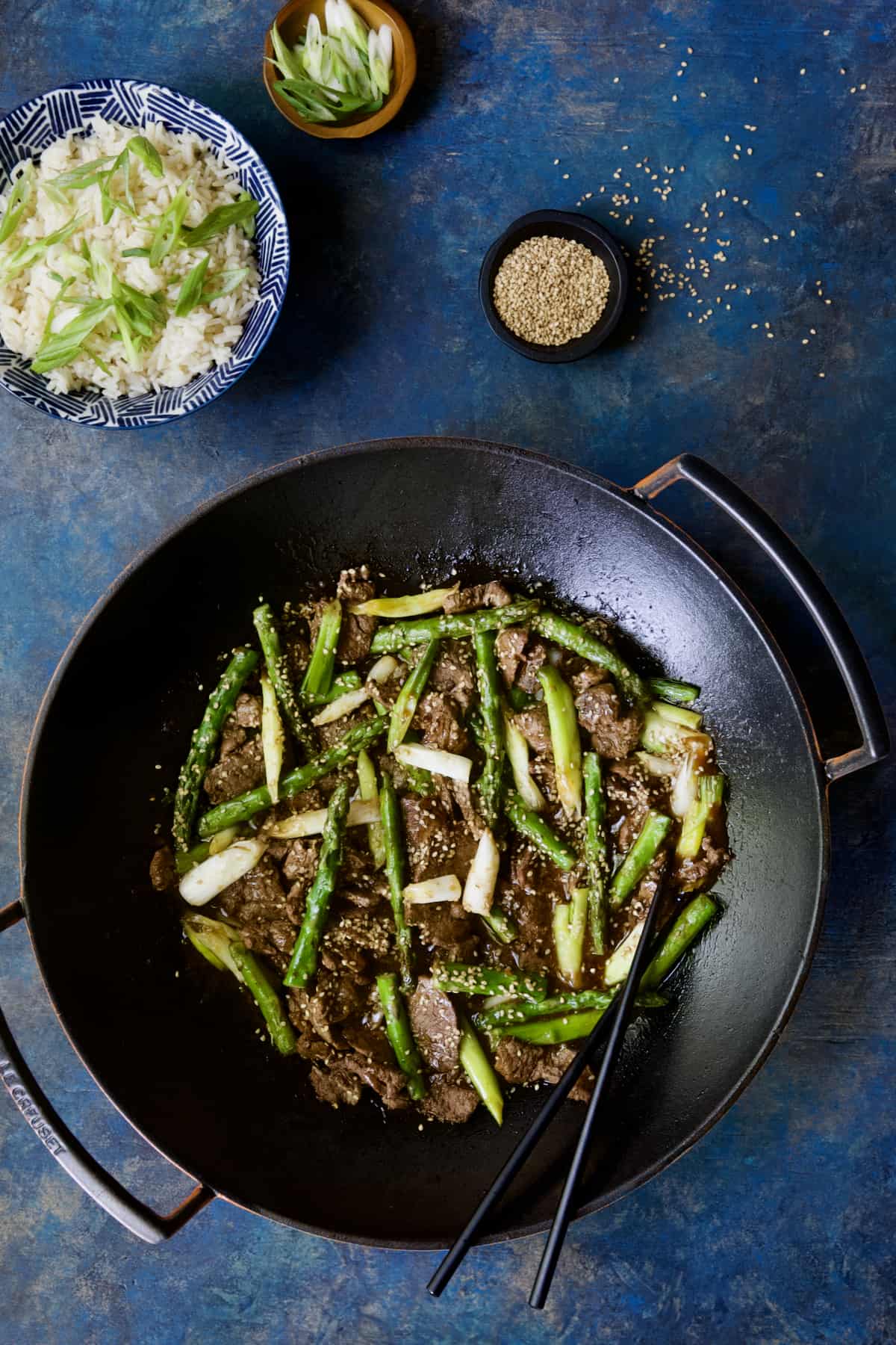 Sesame Beef and Asparagus Stir-Fry in cast iron wok on bright blue background.