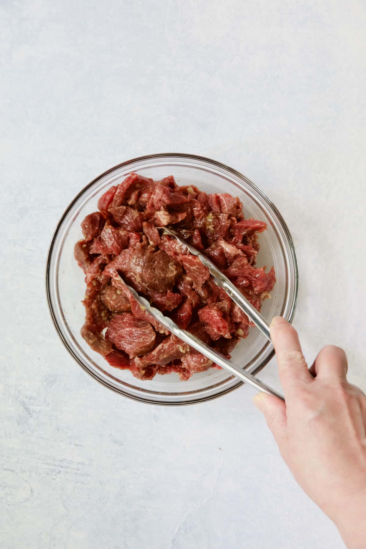 Beef being tossed with tongs in glass bowl.