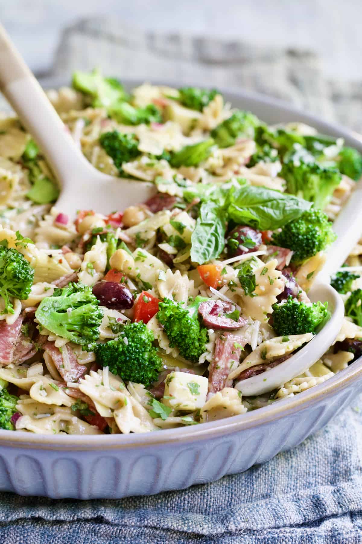 Antipasto Pasta Broccoli Salad with Parmesan Herb Vinaigrette with serving utensils in gray bowl.
