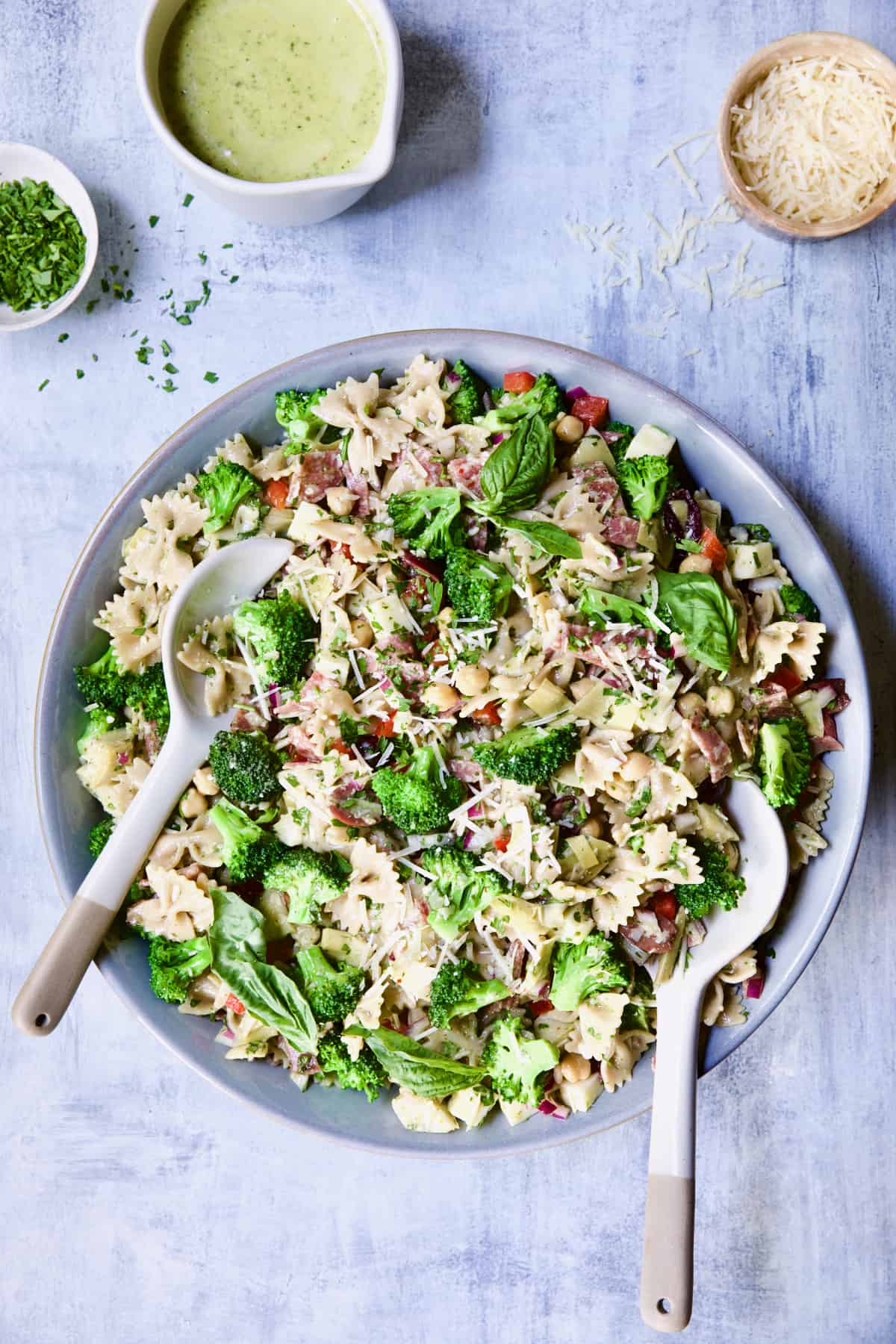 Antipasto Pasta Broccoli Salad with Parmesan Herb Vinaigrette in gray bowl with serving utensils on blue background.