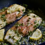 Close-up photo of Prosciutto-Wrapped Halibut with Herb - Caper Butter with lemon wedges in black skillet.