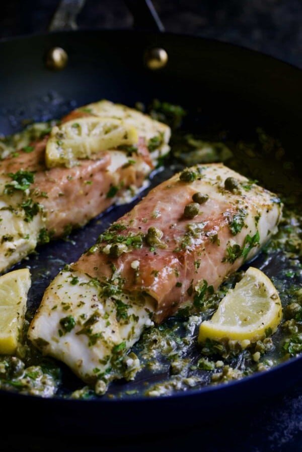 Close-up photo of Prosciutto-Wrapped Halibut with Herb - Caper Butter with lemon wedges in black skillet.