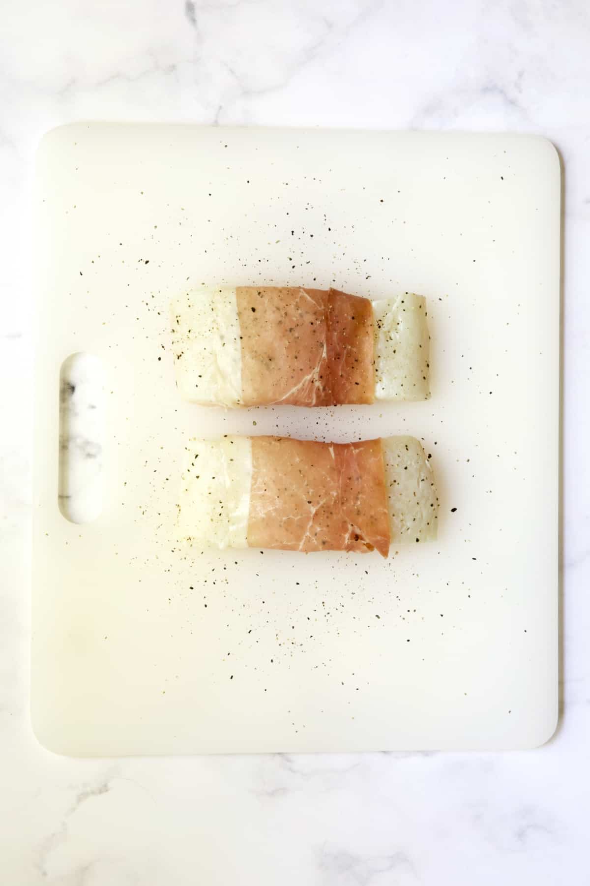 Two prosciutto-wrapped halibut fillets seasoned with pepper on a white plastic cutting board before being cooked.