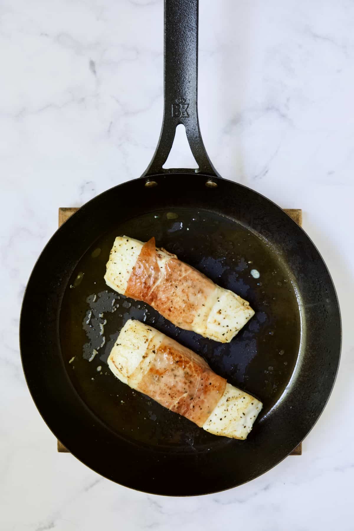 Two prosciutto-wrapped halibut fillets in black skillet being browned.