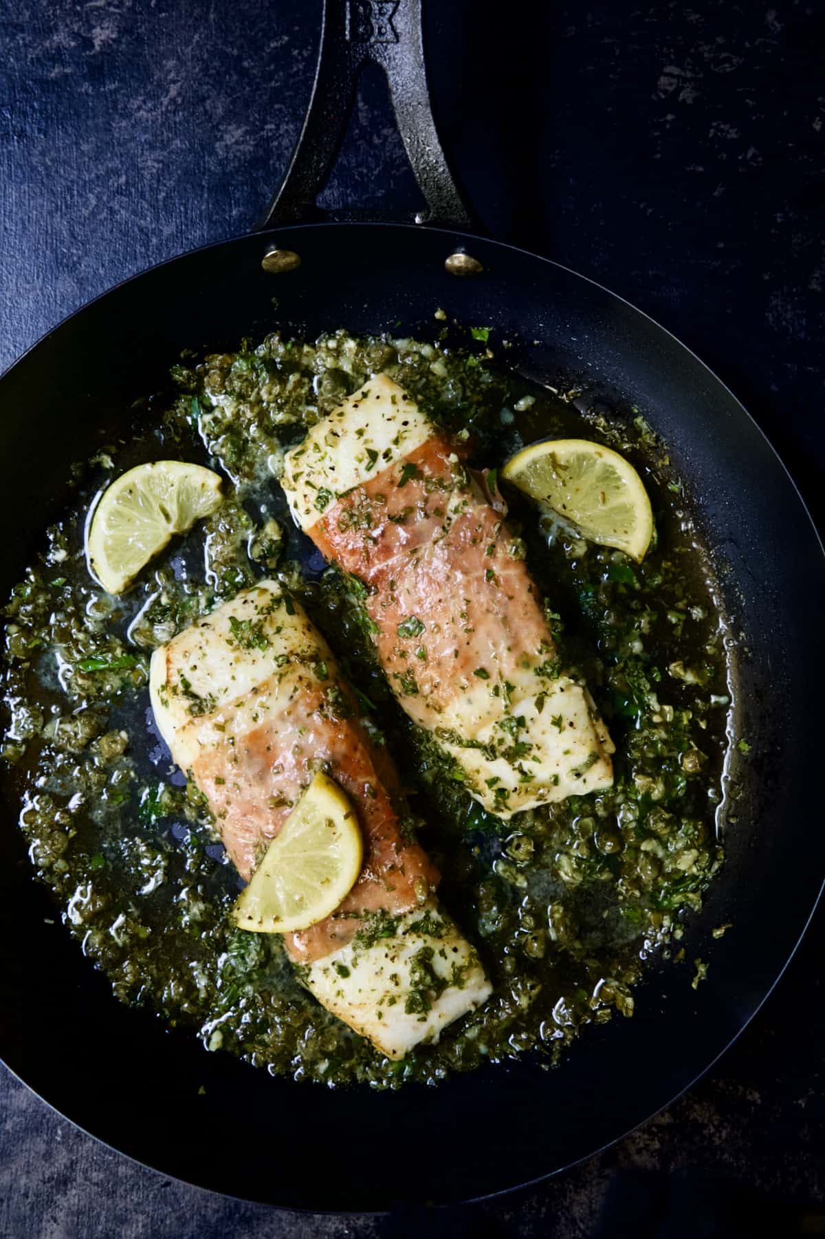 Prosciutto-Wrapped Halibut with Herb - Caper Butter in black skillet with lemon wedges.