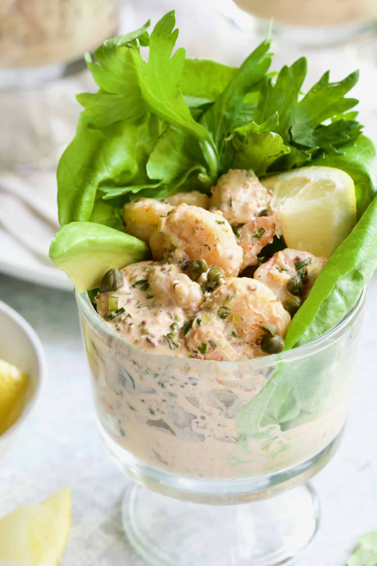 Sriracha Shrimp Remoulade Cocktail in footed glass with avocado, lemon, lettuce and celery sprigs.