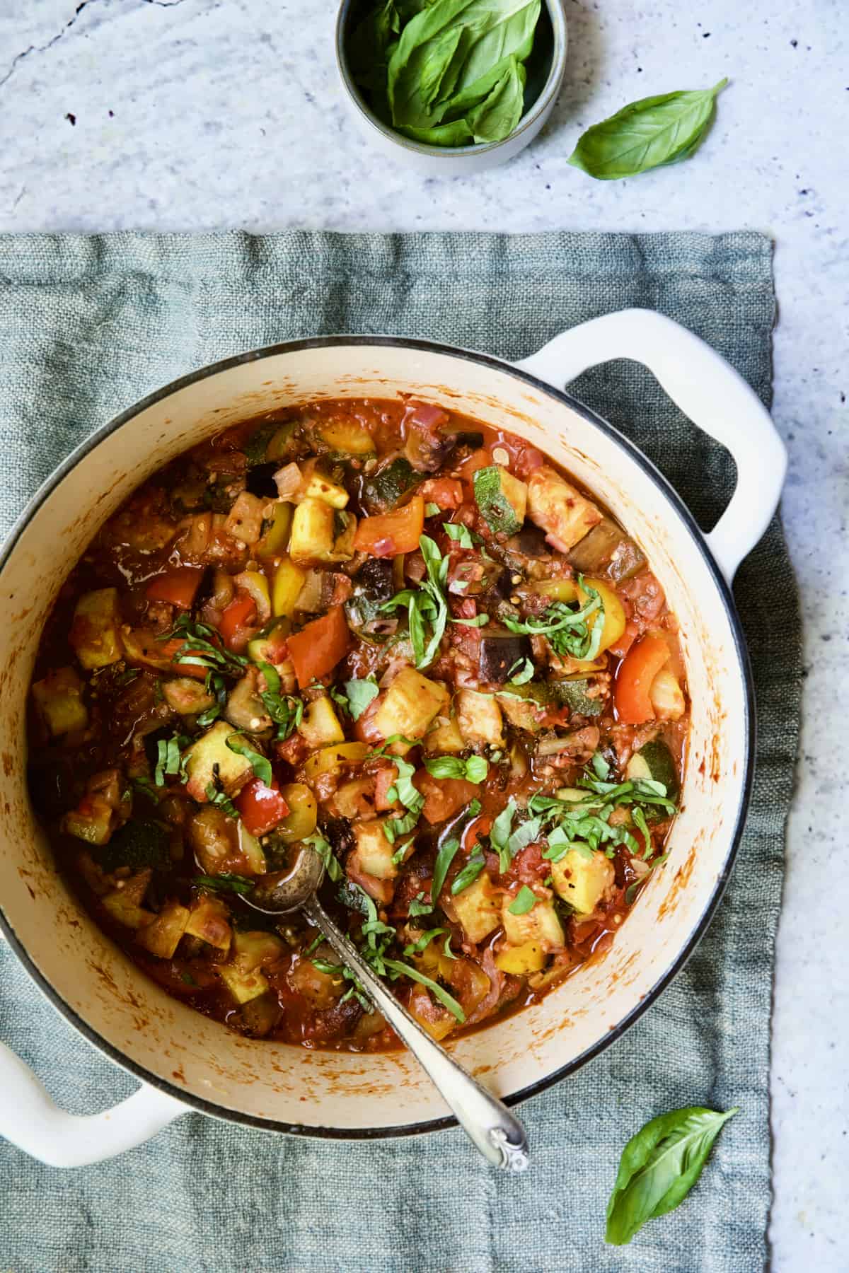 Classic French Ratatouille in white Dutch oven with spoon on blue napkin.