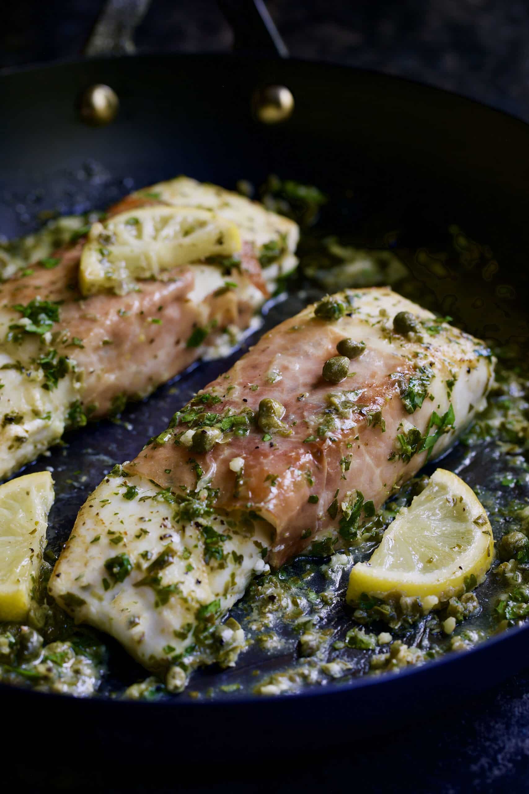 Prosciutto-Wrapped Halibut with Herb - Caper Butter in black skillet with lemon wedges.