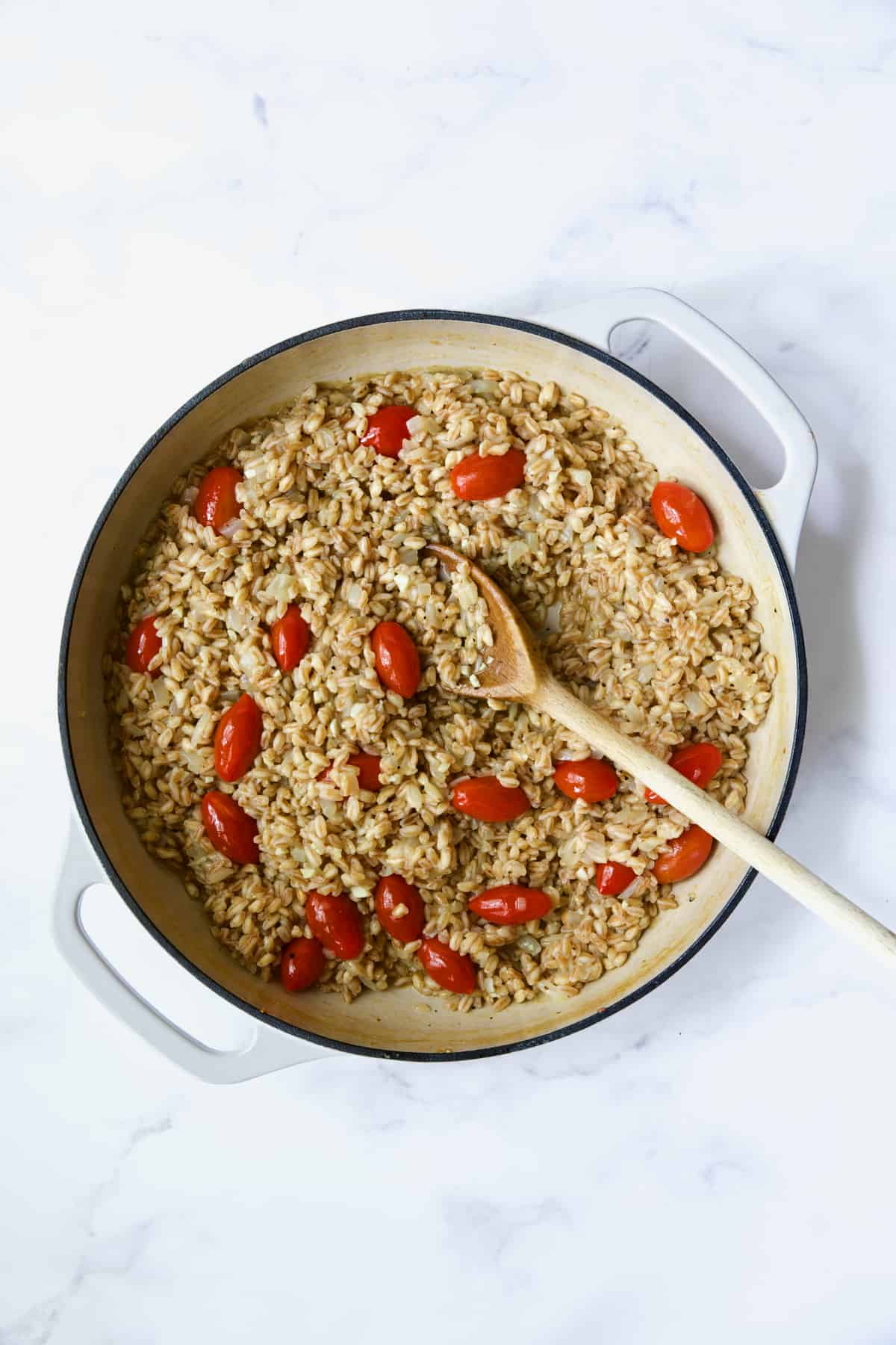 Process photo of farro risotto after tomatoes are added being stirred with wooden spoon.