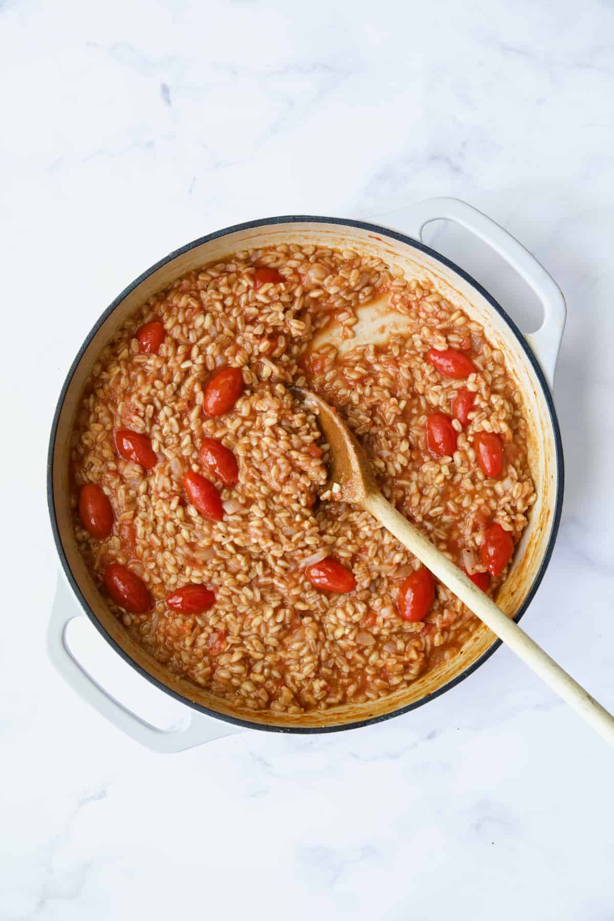 Process photo of farro risotto after crushed tomatoes are added being stirred with wooden spoon.