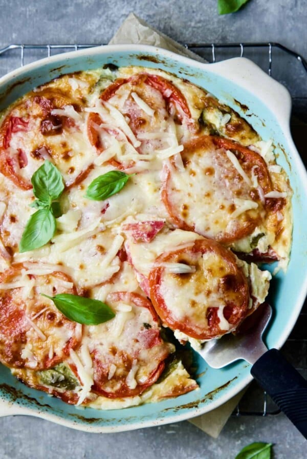Crustless Southern Tomato Pie in light blue pie pan garnished with fresh basil with a piece cut out and about to be served.