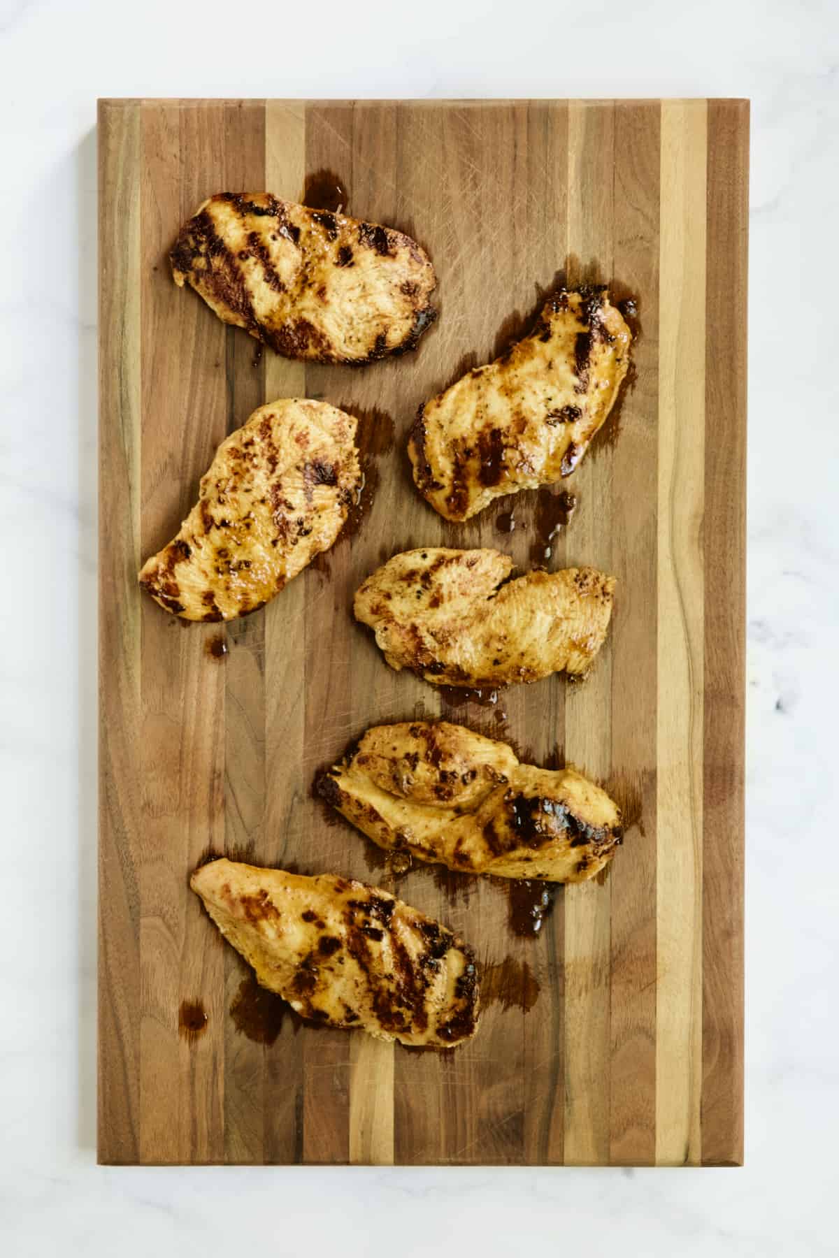 Six pieces of grilled chicken breasts on wood cutting board.