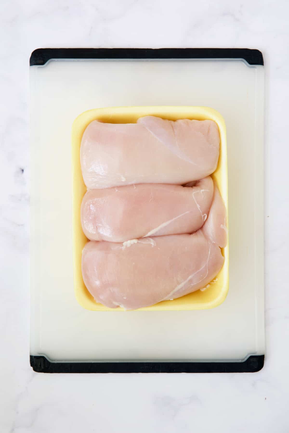 Three chicken breasts in yellow styrofoam container on white plastic cutting board.