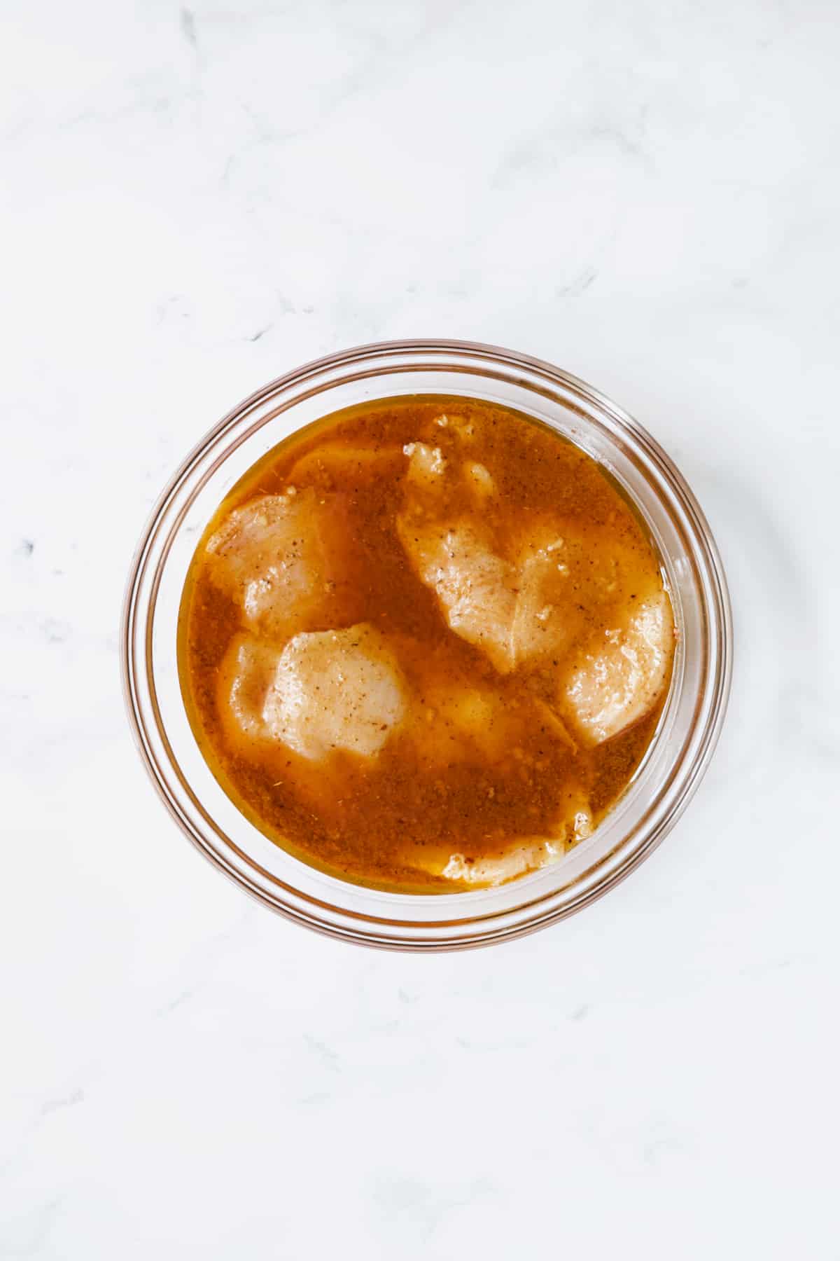 Uncooked chicken breasts in marinade in glass bowl.