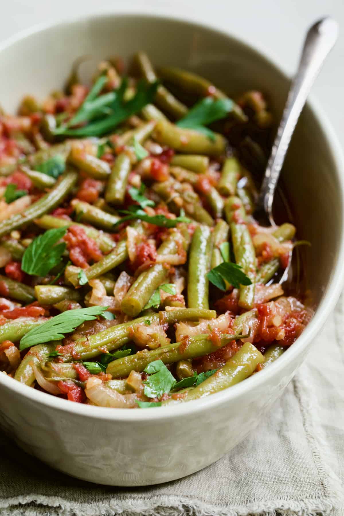 Mediterranean Braised Green Beans with Tomatoes in serving dish with serving spoon on beige fringed napkin.