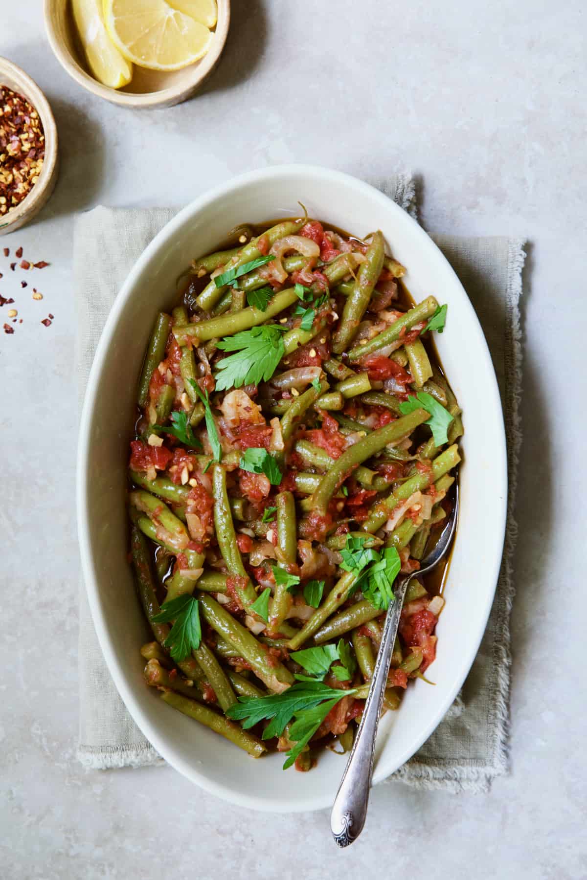 Mediterranean Braised Green Beans with Tomatoes in oval serving dish with serving spoon garnished with fresh parsley.