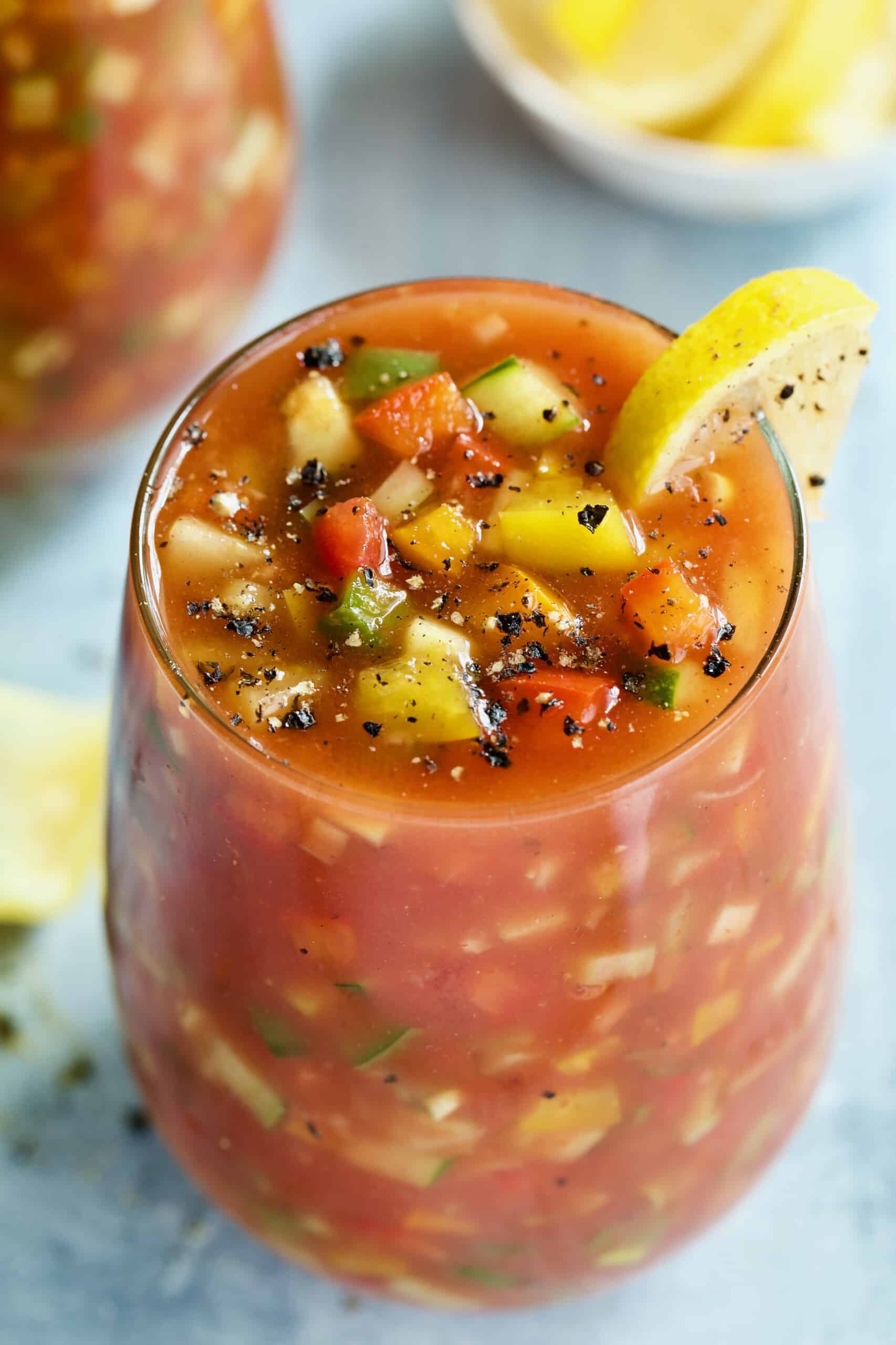 One glass of Best Vegetable Gazpacho on light blue background garnished with lemon wedge.