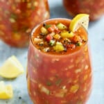 Glass of Very Veggie Gazpacho on light blue background garnished with lemon wedges.