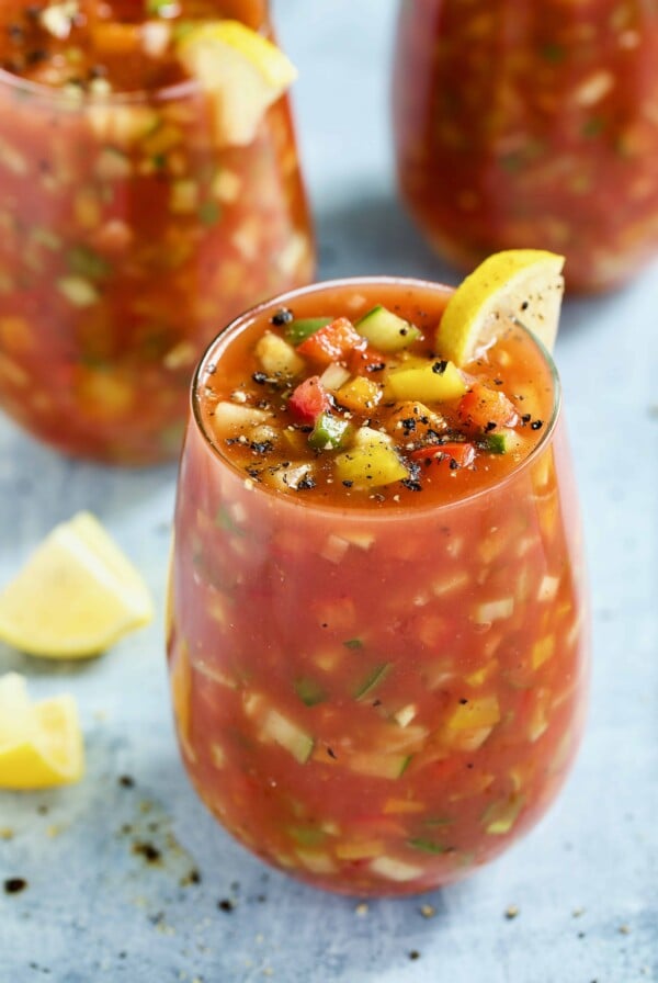 Glass of Very Veggie Gazpacho on light blue background garnished with lemon wedges.