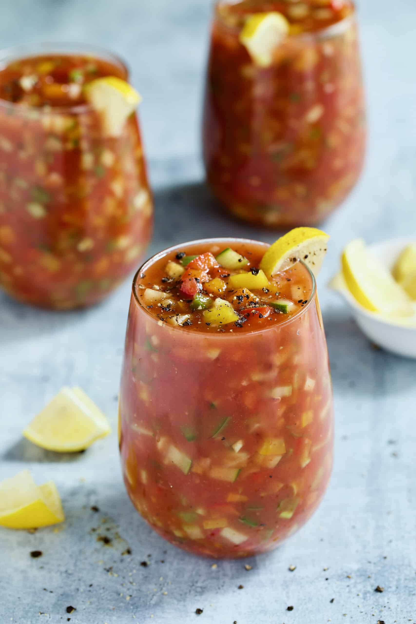 Three glasses of Very Veggie Gazpacho on light blue background garnished with lemon wedges.