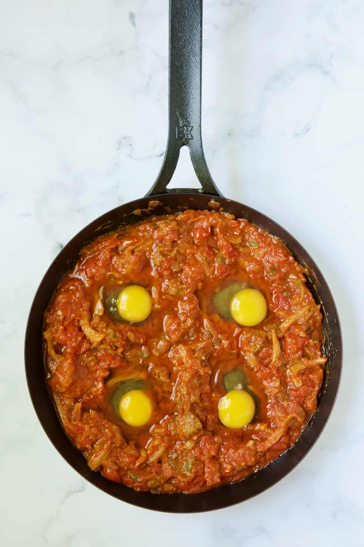 Four uncooked eggs nestled in tomato sauce in skillet.