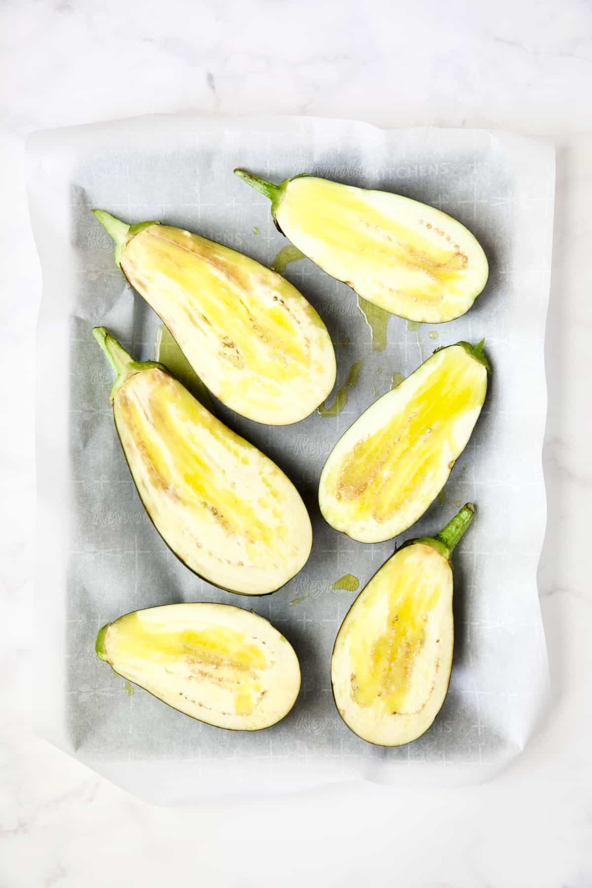 Six eggplant halves on parchment paper-lined baking sheet drizzled with olive oil before being roasted.