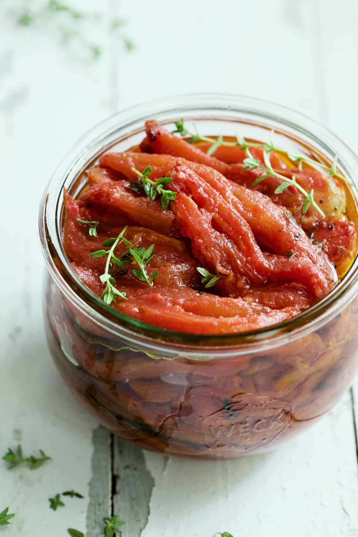 Tomato Confit in glass jar garnished with fresh thyme sprigs on white distressed background.
