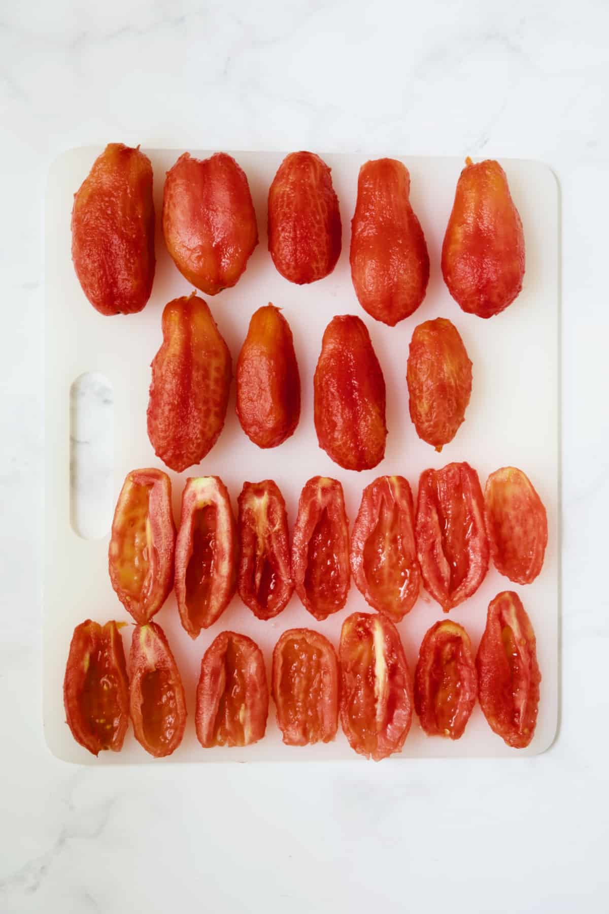 Nine whole skinned tomatoes and 14 peeled, halved and seeded tomatoes on white cutting board.