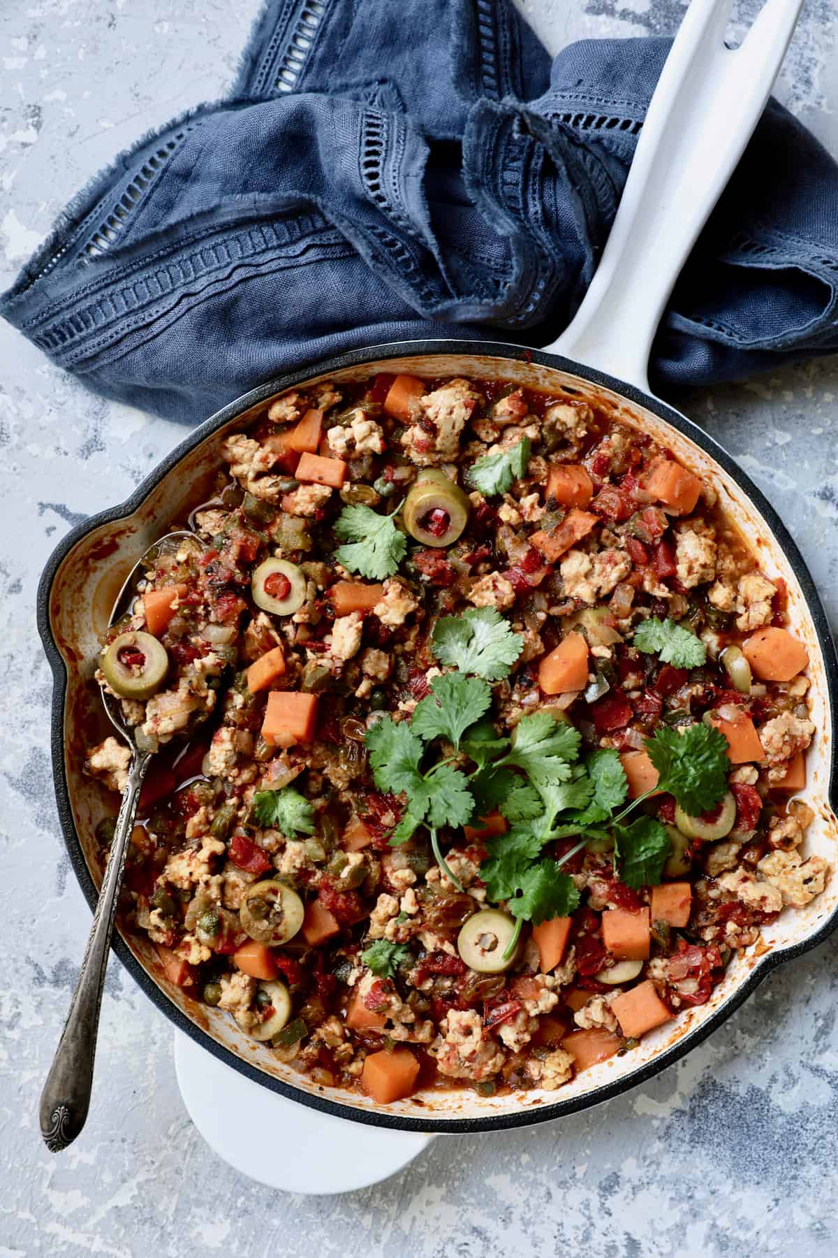 Cuban-Style Turkey Picadillo with Sweet Potatoes in white cast iron enameled skillet on blue background with blue napkin.